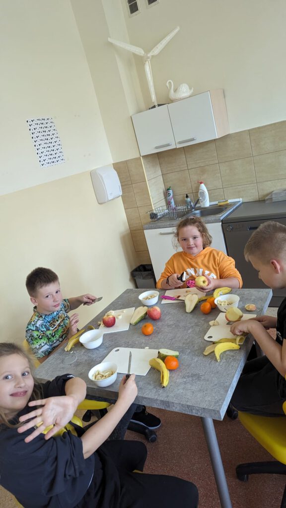 Dzieci siedzą przy stoliku i przygotowują sałatkę z owoców.
